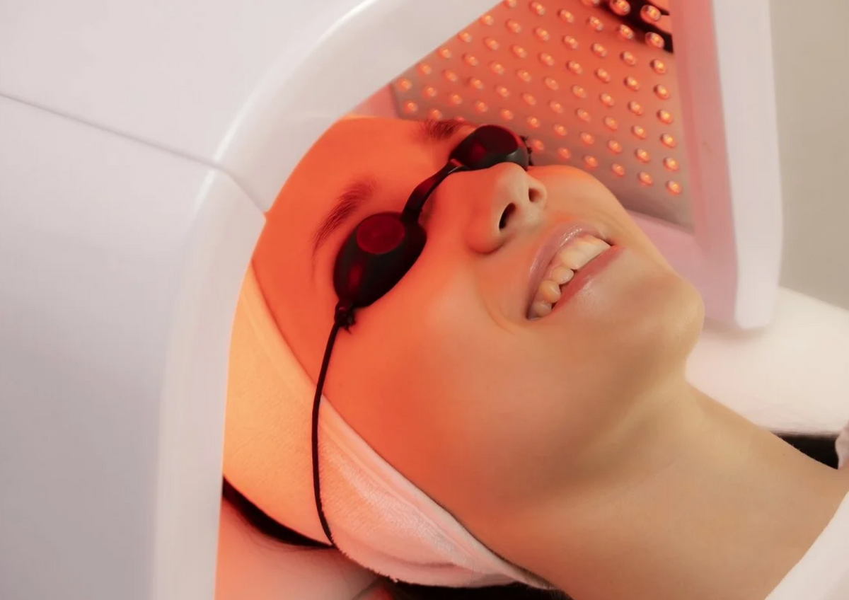 Mujer con gafas protectoras durante una sesión de fototerapia facial bajo un panel de luz LED roja.