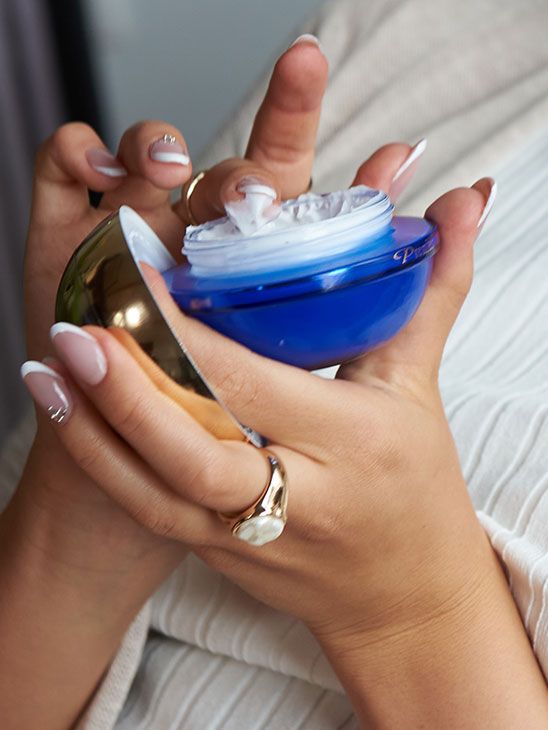Manos sosteniendo un tarro azul abierto de crema para el contorno de ojos Premier Prestige con textura blanca.