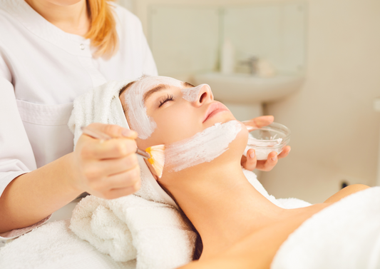 Aplicación de mascarilla facial blanca con pincel en el mentón durante el tratamiento de firmeza y elasticidad.