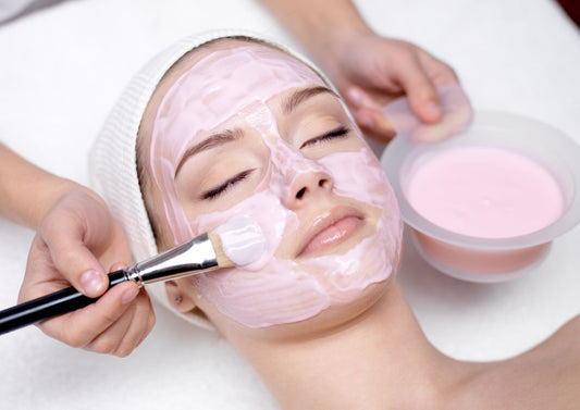 Aplicación de mascarilla facial rosa con pincel sobre el rostro de una mujer durante una higiene profunda.